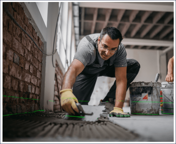 A man wearing gloves and glasses is laying tile adhesive on a floor in an unfinished room with brick walls.